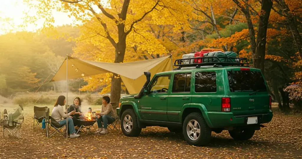 秋の紅葉が美しいキャンプ場で、Jeepコマンダー（グリーン）の横にタープが張られ、家族がBBQを楽しんでいる様子。車にはルーフラックが装着され、アウトドアギアが満載。暖かくアクティブな雰囲気の写真